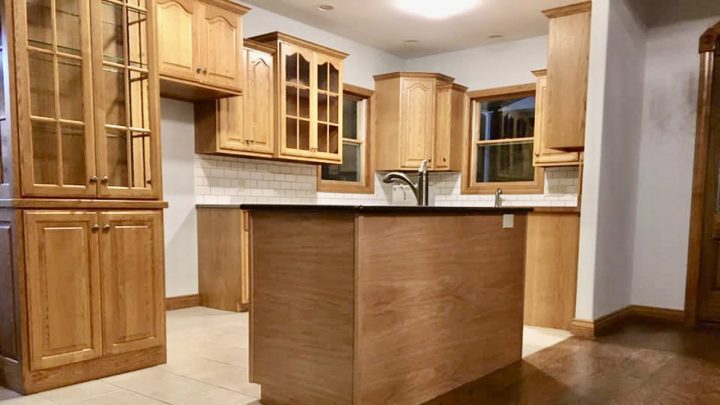 A modern kitchen with light wooden cabinets, some of which have glass fronts. The backsplash is made of white subway tiles, and there is a central island with a dark countertop. The floor is tiled, and the room has a clean, organized appearance.