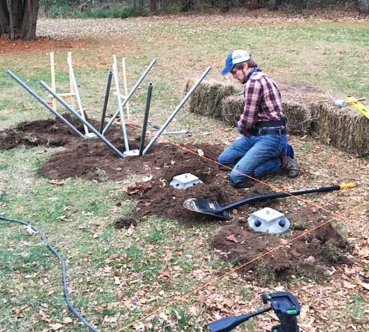 A man kneeling on the ground while installing metal poles into the soil. He is wearing a plaid shirt and jeans, and there are tools and materials scattered around him. The area is surrounded by grass and hay bales.