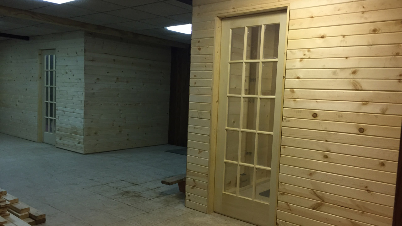 An unfinished interior space featuring light wooden walls and a glass-paneled door. The room has a concrete floor, exposed ceiling beams, and some construction materials scattered around.