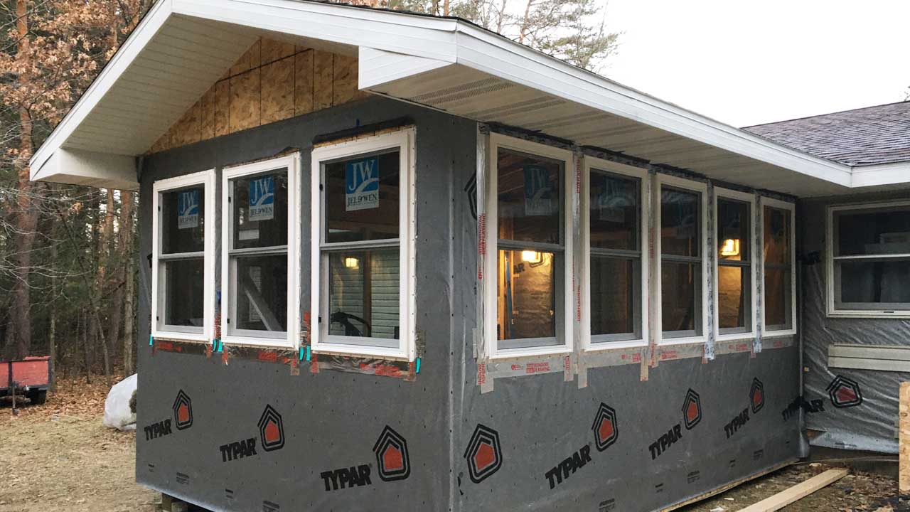 A partially constructed home addition with gray exterior insulation and finish system (EIFS) panels. The windows are installed, and the structure is surrounded by trees in a wooded area.