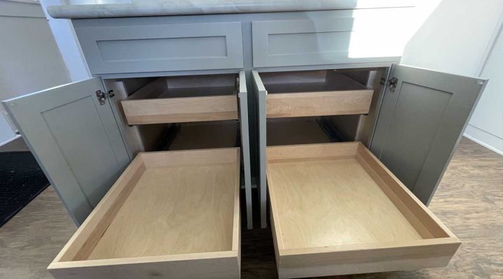 A close-up view of open cabinets and drawers in a kitchen. The cabinets are painted gray, and the drawers are made of light wood. The space appears clean and organized, with no items inside the drawers.