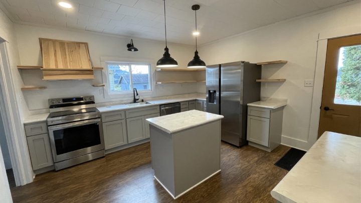 A contemporary kitchen featuring stainless steel appliances, including a refrigerator and oven. The kitchen has white cabinets, a central island with a marble countertop, and hardwood flooring. Pendant lights hang from the ceiling.