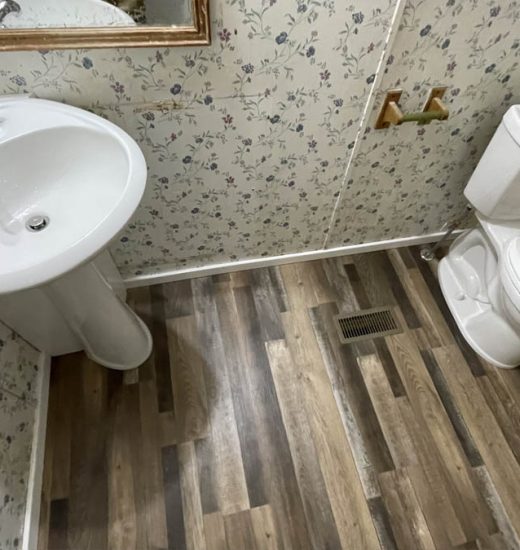 A bathroom featuring floral wallpaper, a white pedestal sink, and a toilet. The floor is covered with wood-like laminate, and there is a mirror above the sink.