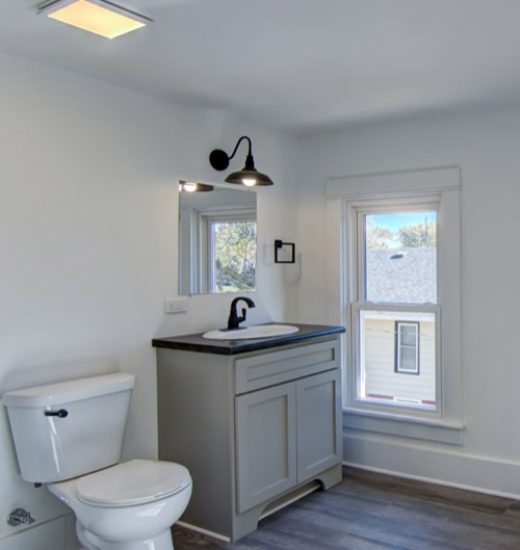 A clean and modern bathroom with white walls and a gray vanity cabinet. The room includes a toilet, a sink with a black faucet, and a window that lets in natural light. The floor is covered with wood-like laminate.