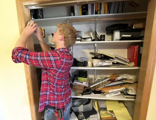 A man wearing a red plaid shirt is using a drill to install a light fixture on the ceiling of a cluttered closet. The shelves are filled with books, papers, and various items.