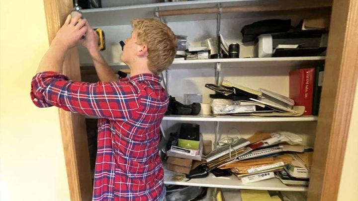 A man wearing a red plaid shirt is using a drill to install a light fixture on the ceiling of a cluttered closet. The shelves are filled with books, papers, and various items.