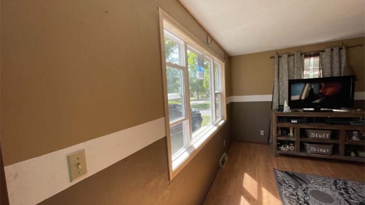 A living room with brown walls and a white trim. The room features a large window with a view outside, a flat-screen TV on a wooden stand, and a patterned rug on the wooden floor. The space is well-lit and cozy.