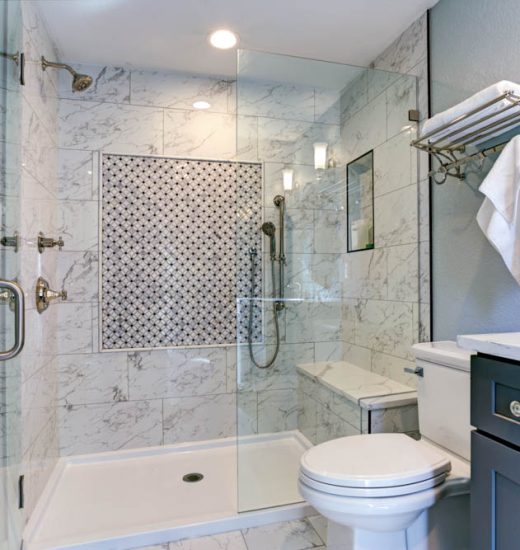 A luxurious bathroom with marble tiles on the walls and floor. It includes a walk-in shower with a glass door, a toilet, and a blue vanity cabinet. There are white towels neatly hung on racks, and the lighting is soft and warm.