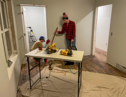 Two workers are renovating a room. One worker is using a circular saw on a table, while the other holds a broom. The room has light-colored walls and wooden flooring, and various tools and materials are scattered around.