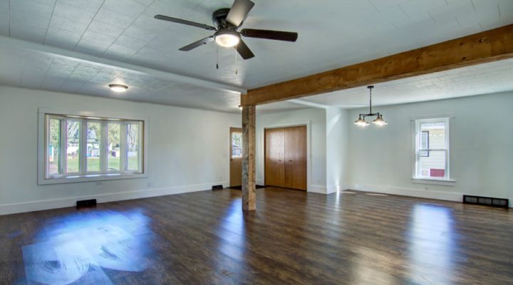 A large, empty living room with white walls and a wooden floor. The room features a ceiling fan, recessed lighting, and a prominent wooden beam running across the ceiling. There are two windows allowing natural light to enter.