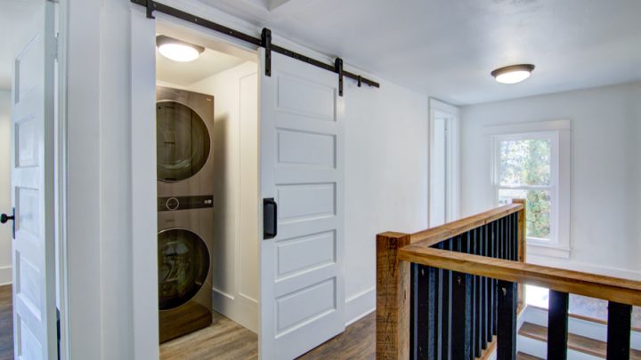 A laundry room with a sliding barn door, revealing a washer and dryer inside. The room has white walls, wooden flooring, and a staircase with a wooden railing leading to another level. The space is well-lit with ceiling lights.