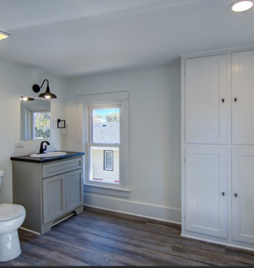 A clean and bright bathroom with white cabinets, a sink vanity, and a toilet. The room has a window that allows natural light to enter, and there is a storage cabinet for extra supplies. The floor is made of wood-like laminate.