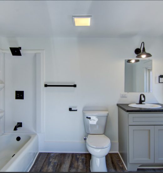 : A small but well-organized bathroom with white walls and a wooden floor. It features a bathtub with a shower curtain, a toilet, and a gray vanity cabinet with a black countertop. There is a window letting in natural light.