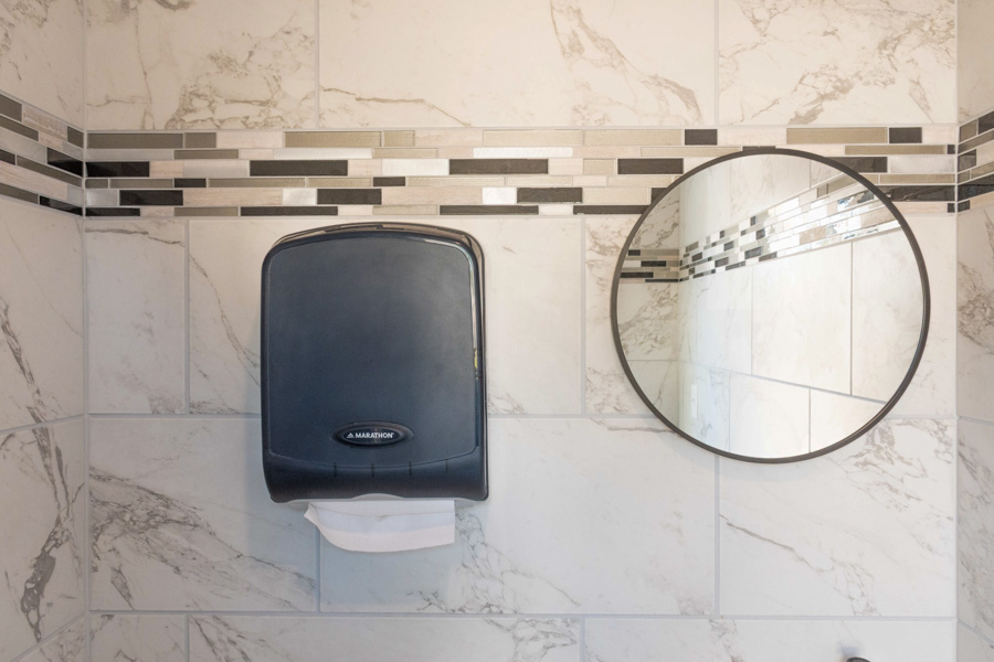 A clean, commercial-style bathroom wall with a black paper towel dispenser, round mirror, and marble-look tile accented by a mosaic tile band.